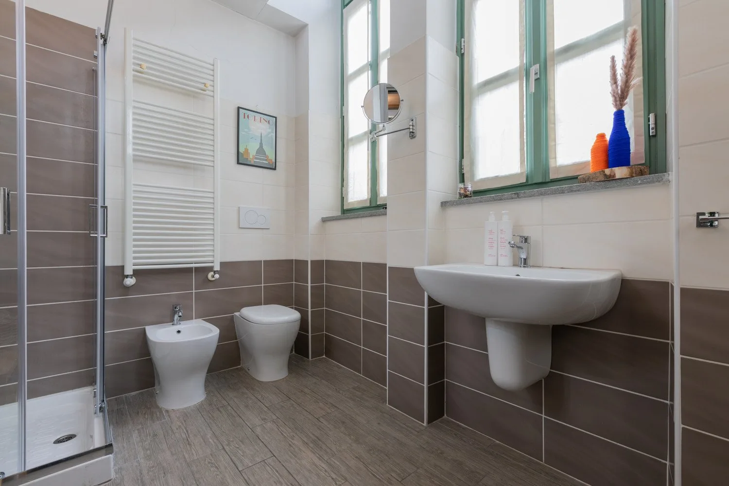 Un baño alicatado con ducha de cristal, inodoro y lavabo bajo una ventana.