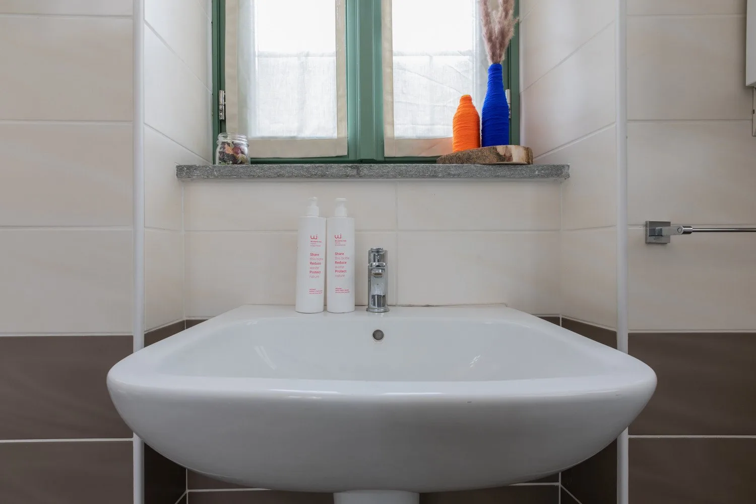 Un lavabo de baño está bajo una ventana con marco verde y artículos de aseo en el alféizar.