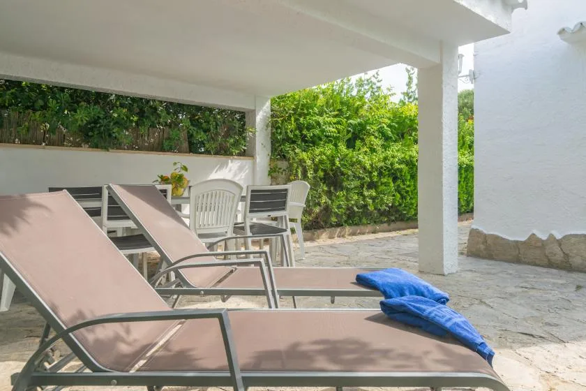 Un patio exterior cubierto tiene asientos reclinables, una mesa y vegetación frondosa.