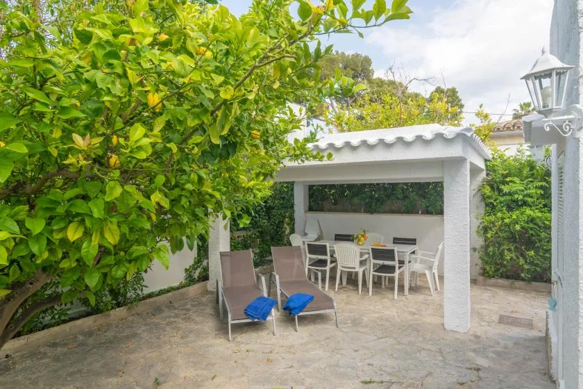 An outdoor dining area sits beneath a covered patio beside leafy trees.
