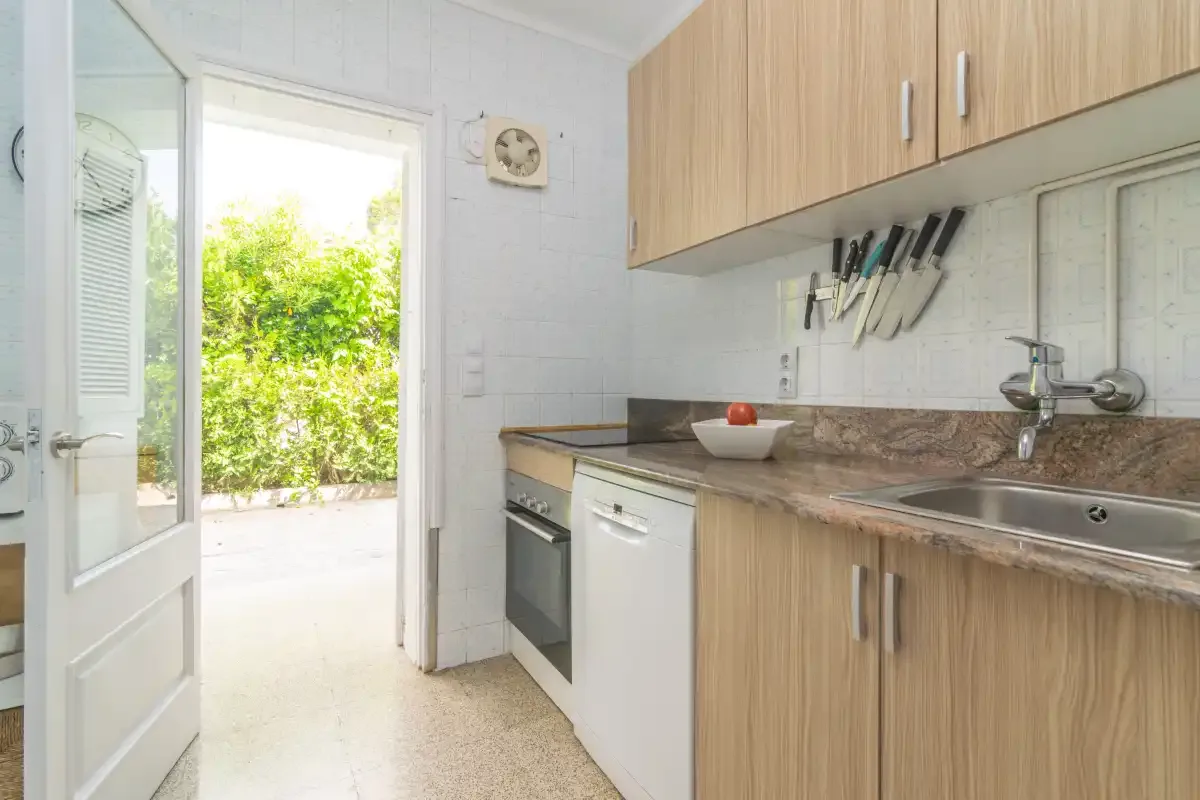 Una cocina con gabinetes de madera, un fregadero y una puerta abierta hacia la vegetación.