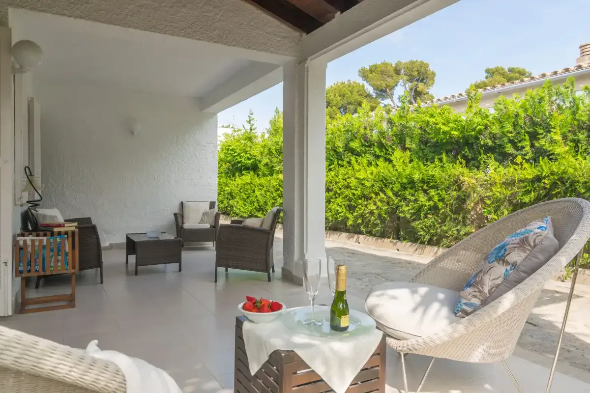 Un patio cubierto con asientos y una mesa pequeña da a un jardín verde.