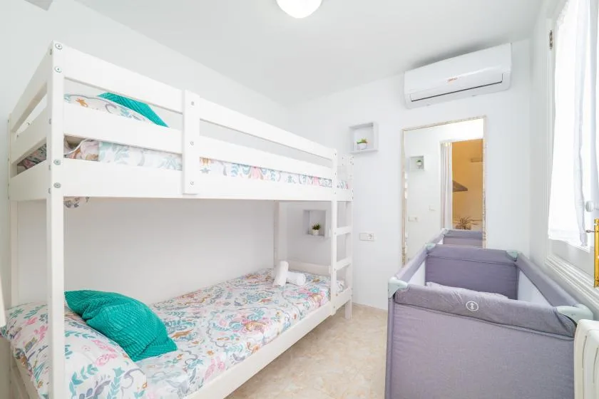 Un dormitorio luminoso con literas blancas y una cuna gris junto a la ventana.