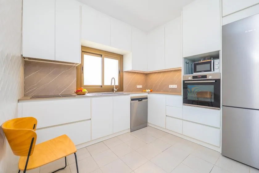 Una cocina blanca y luminosa con ventana, encimera y electrodomésticos integrados.