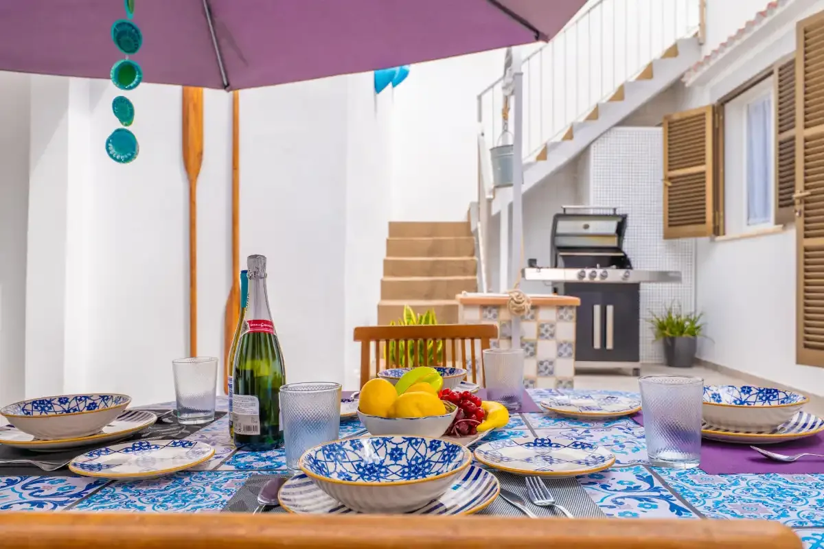 Una mesa de comedor al aire libre bajo una sombrilla morada, con una escalera y una parrilla detrás.