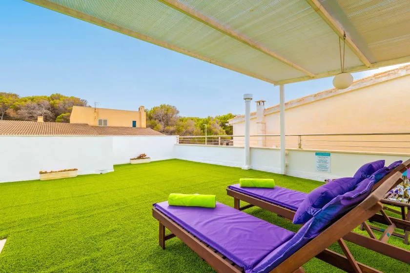 Una terraza en la azotea tiene tumbonas con cojines morados sobre césped artificial verde, bajo un toldo.