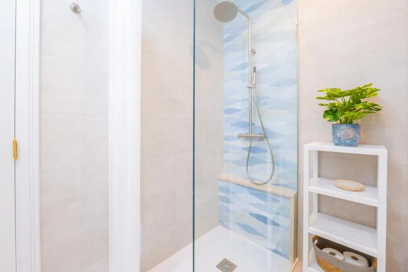 Un baño luminoso cuenta con una ducha de vidrio con azulejos azules y una pequeña estantería.