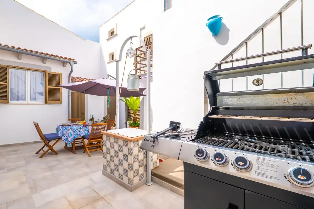 Un patio exterior tiene una mesa de comedor bajo una sombrilla y una parrilla incorporada.