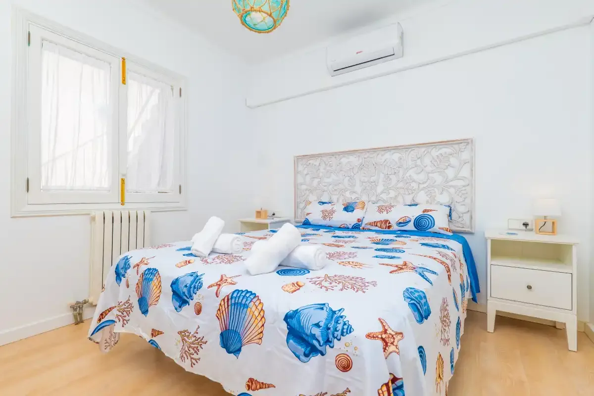 Un dormitorio luminoso con una cama cubierta de motivos de conchas azules y naranjas.