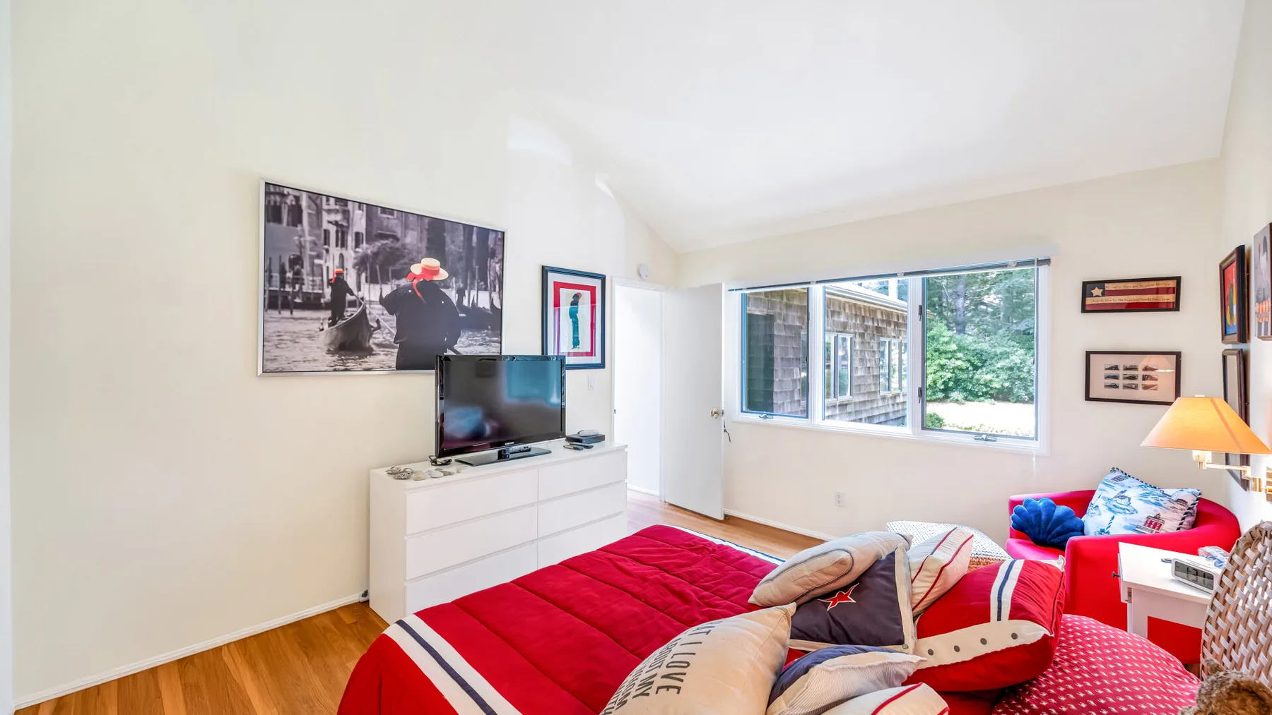 Un dormitorio luminoso tiene una cama con cubierta roja y una cómoda bajo un televisor.