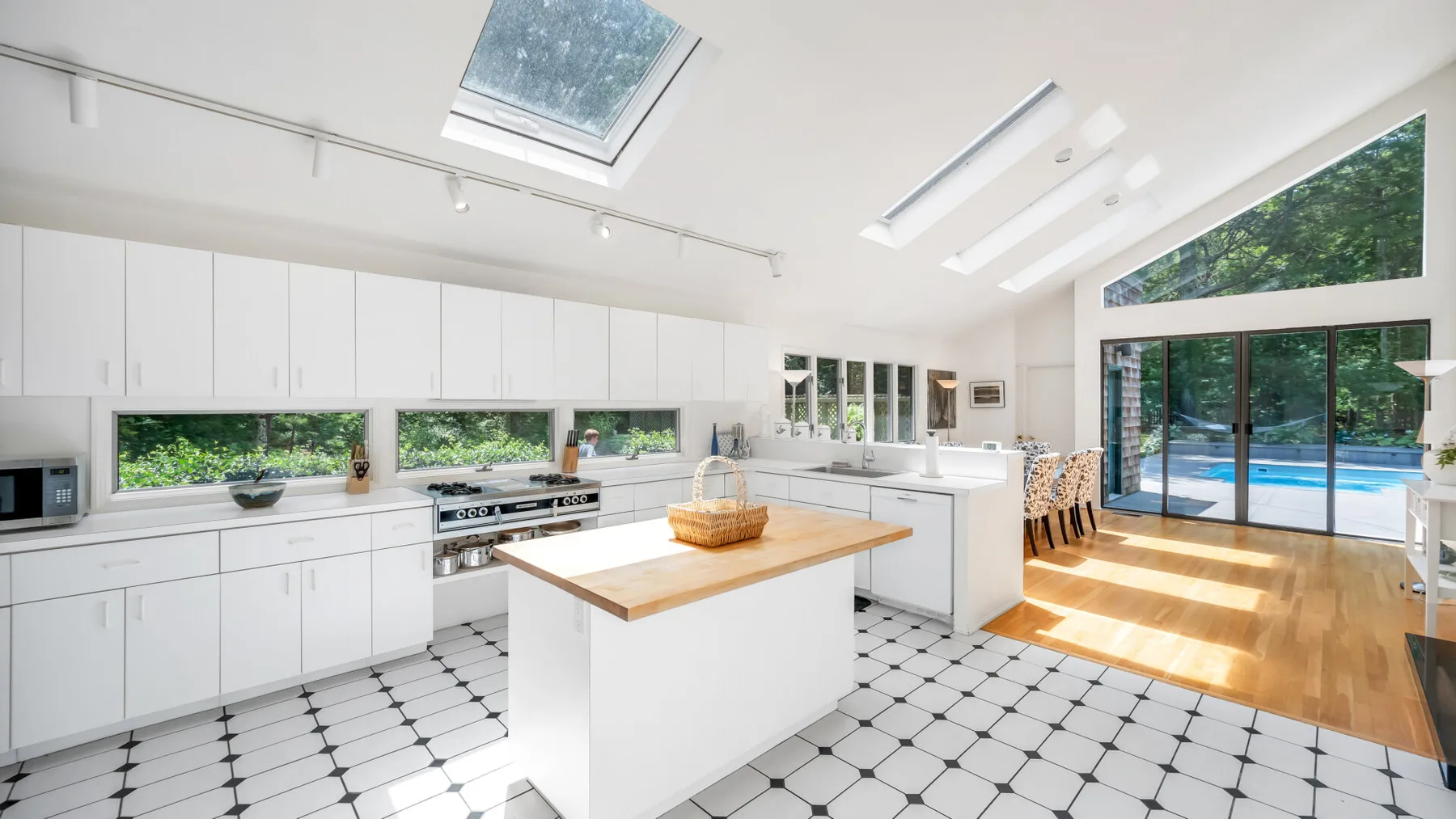 Una cocina blanca y luminosa con una isla central y grandes ventanales con vistas a una piscina.