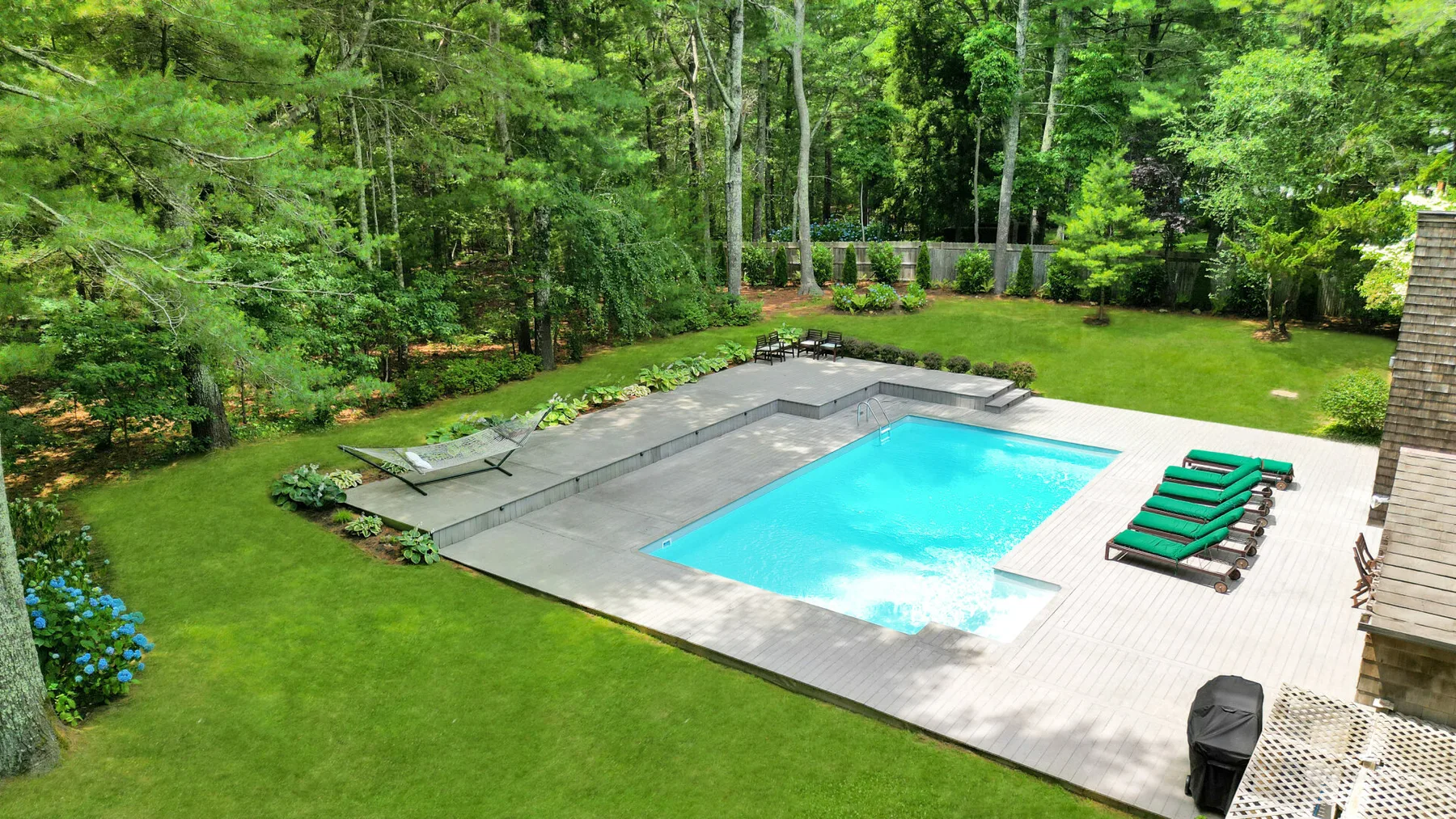 Una piscina al aire libre está en un patio pavimentado rodeado de césped y árboles.