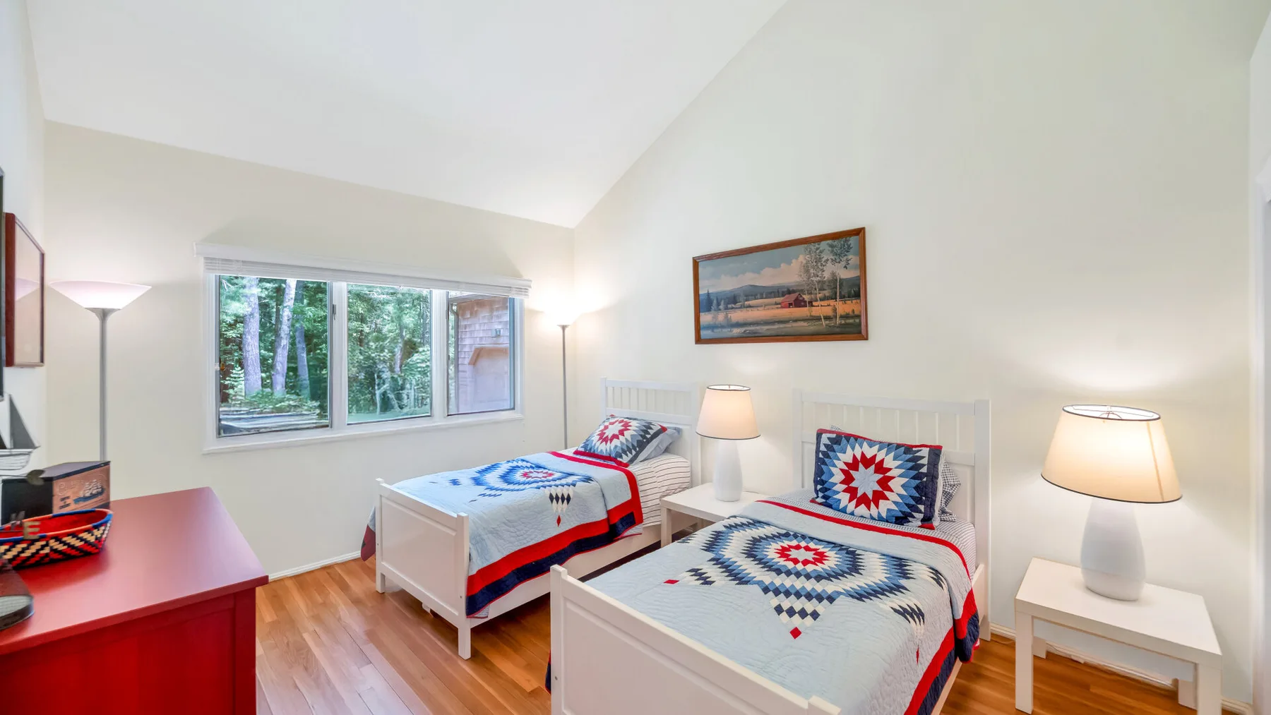 Un dormitorio luminoso con dos camas, ropa de cama colorida y una ventana con vista a los árboles.