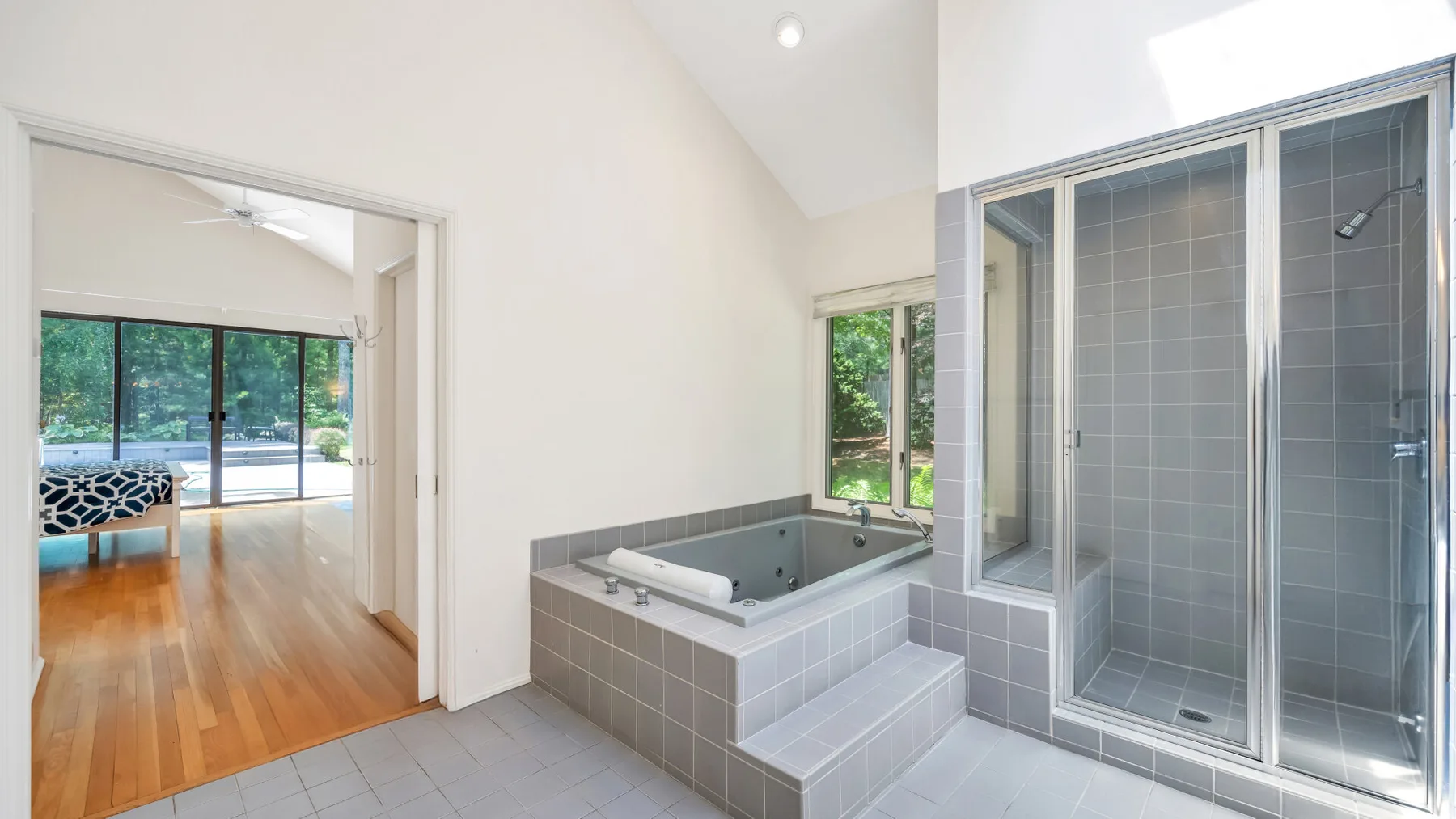 Un baño luminoso con bañera revestida de azulejos y ducha de vidrio junto a una puerta abierta.