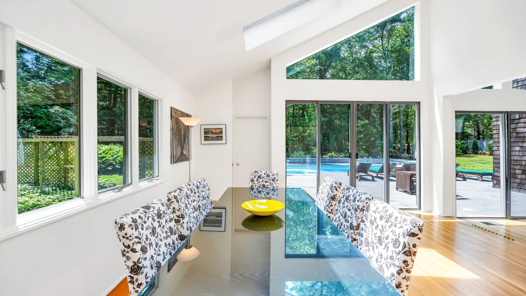 Un comedor luminoso con una mesa de vidrio junto a grandes ventanas que dan a una piscina.