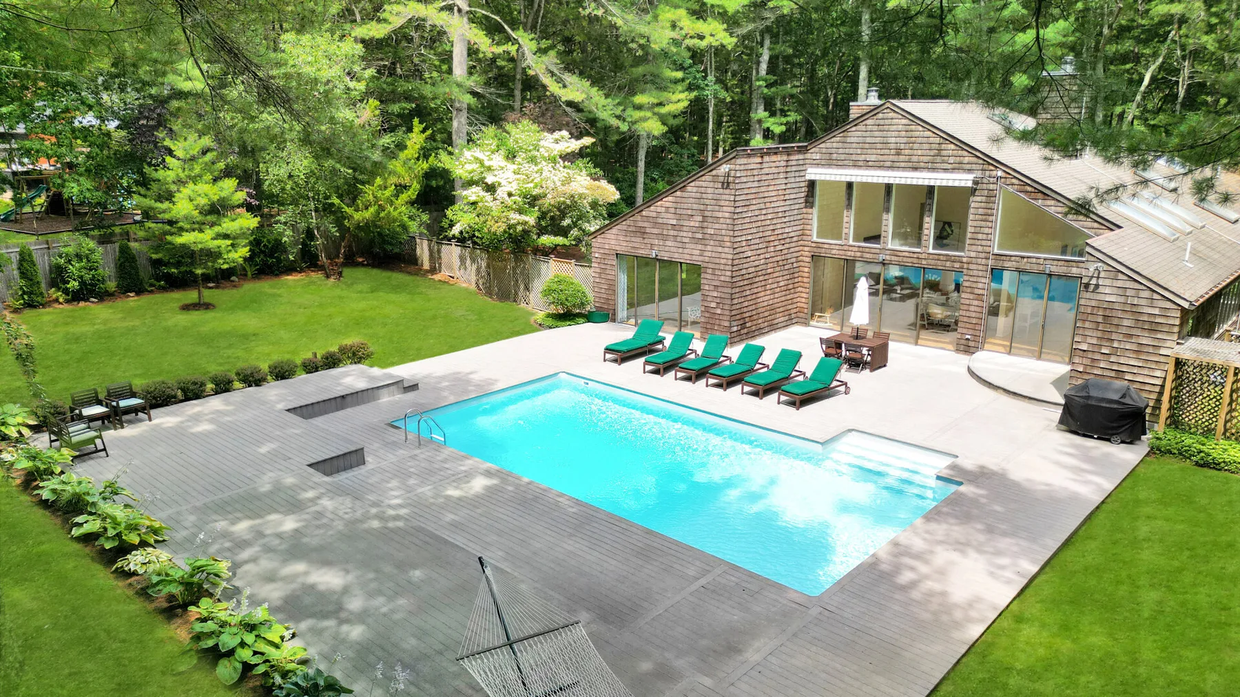 Una piscina exterior y un patio están junto a un edificio revestido de madera y zonas verdes.