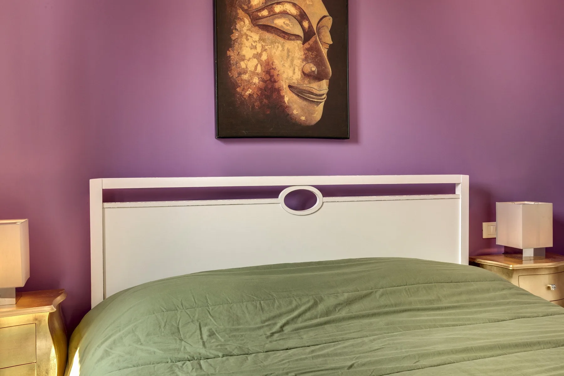 Un dormitorio con una cama bajo un retrato enmarcado sobre una pared morada.