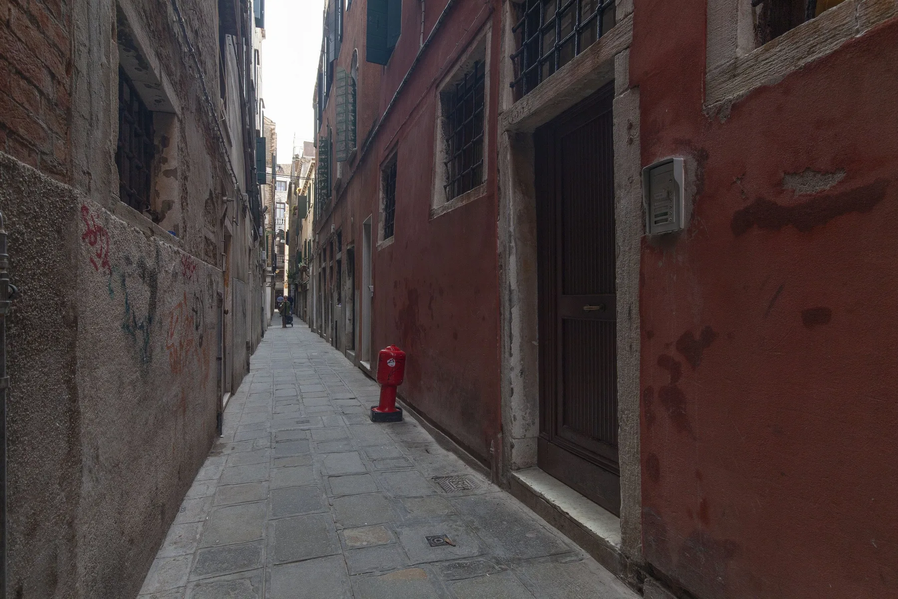 Un callejón estrecho pavimentado pasa entre edificios muy juntos, con un buzón rojo junto a la pared.