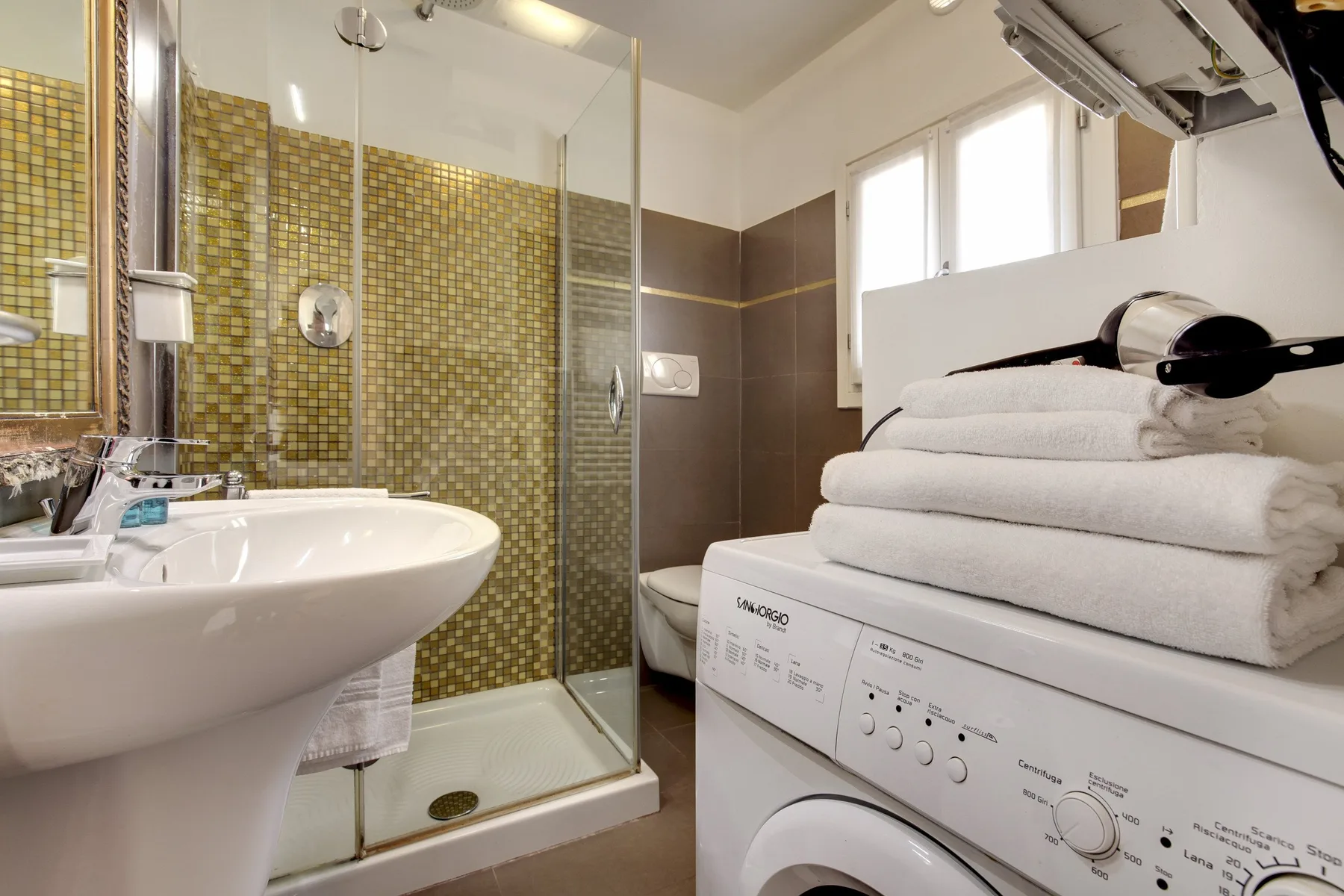 Un baño con ducha de vidrio, lavabo y toallas dobladas sobre una lavadora.