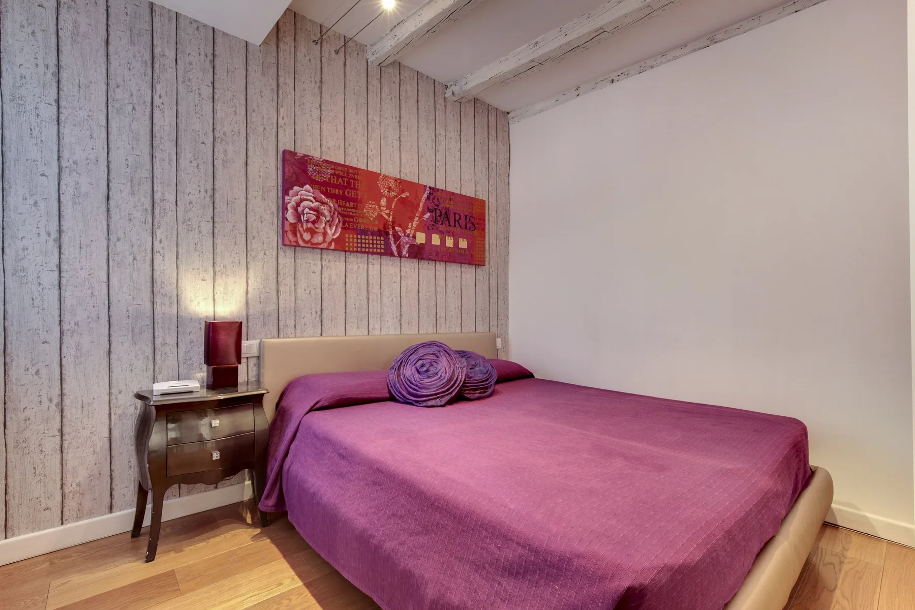 Un dormitorio con una cama cubierta de ropa de cama morada y un cuadro en la pared.
