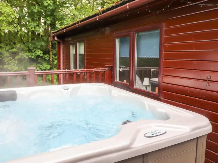 Outdoor hot tub beside a red wooden cabin, surrounded by leafy trees.