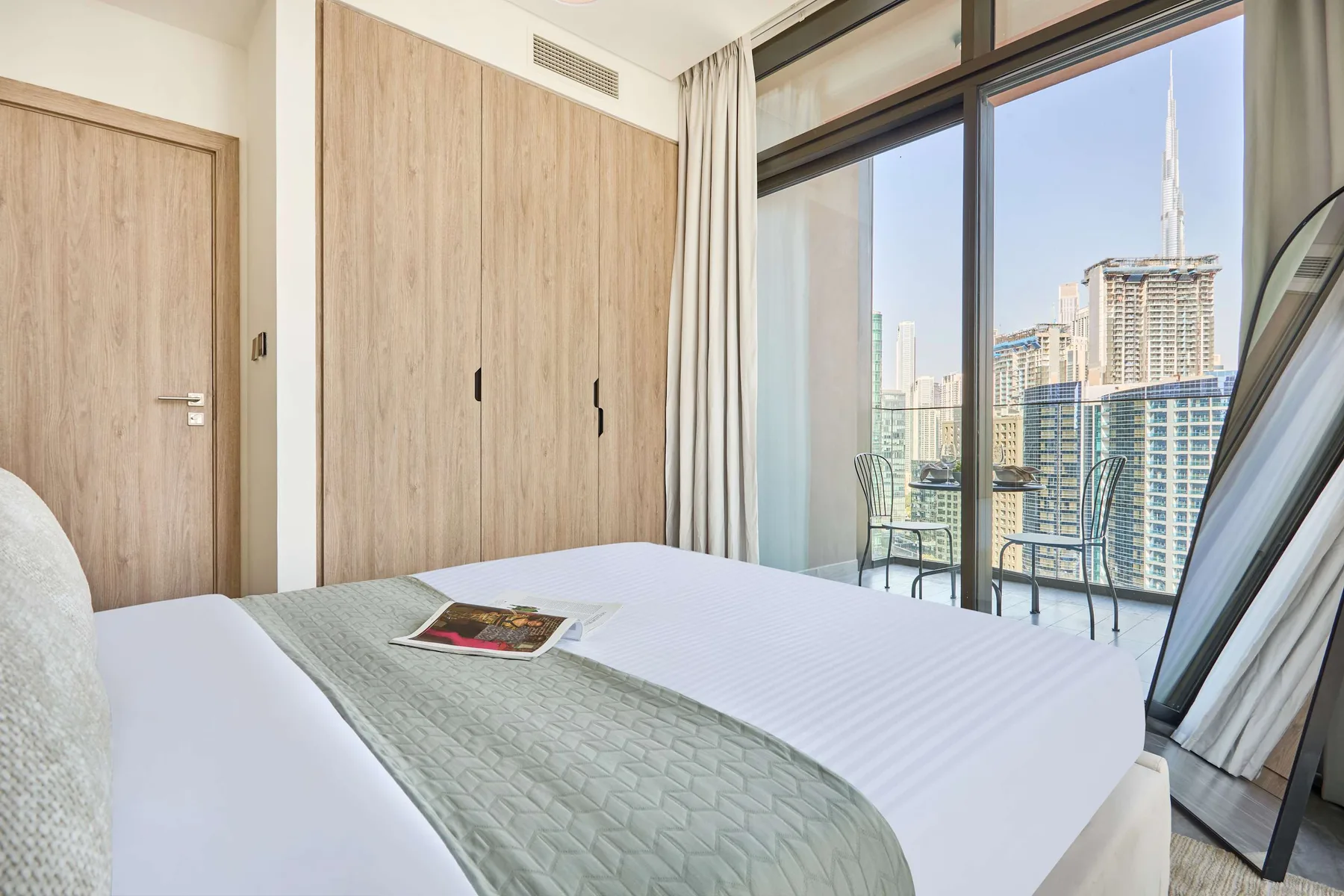 A bedroom with a neatly made bed beside floor-to-ceiling windows overlooking the city.