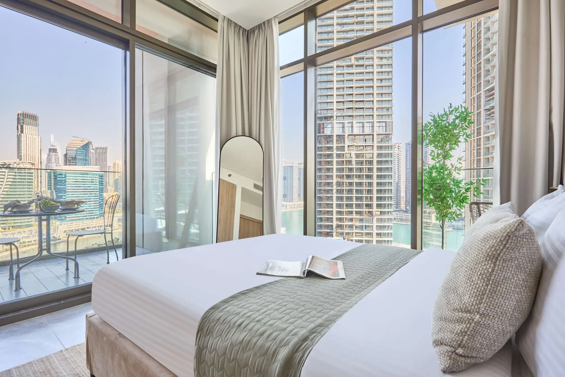 A bedroom with a neatly made bed beside floor-to-ceiling windows overlooking city buildings.