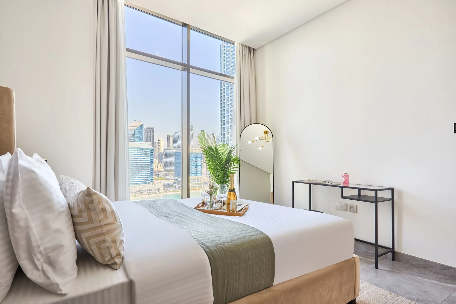 A bright bedroom with a bed beside a large window overlooking city buildings.