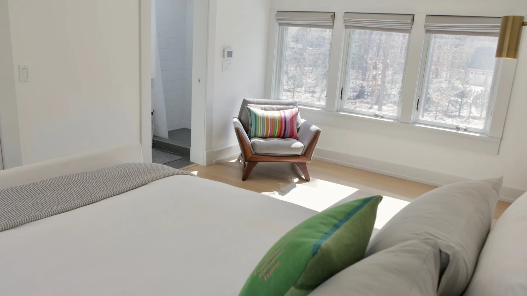 A bright bedroom has a bed and an armchair beside three windows.
