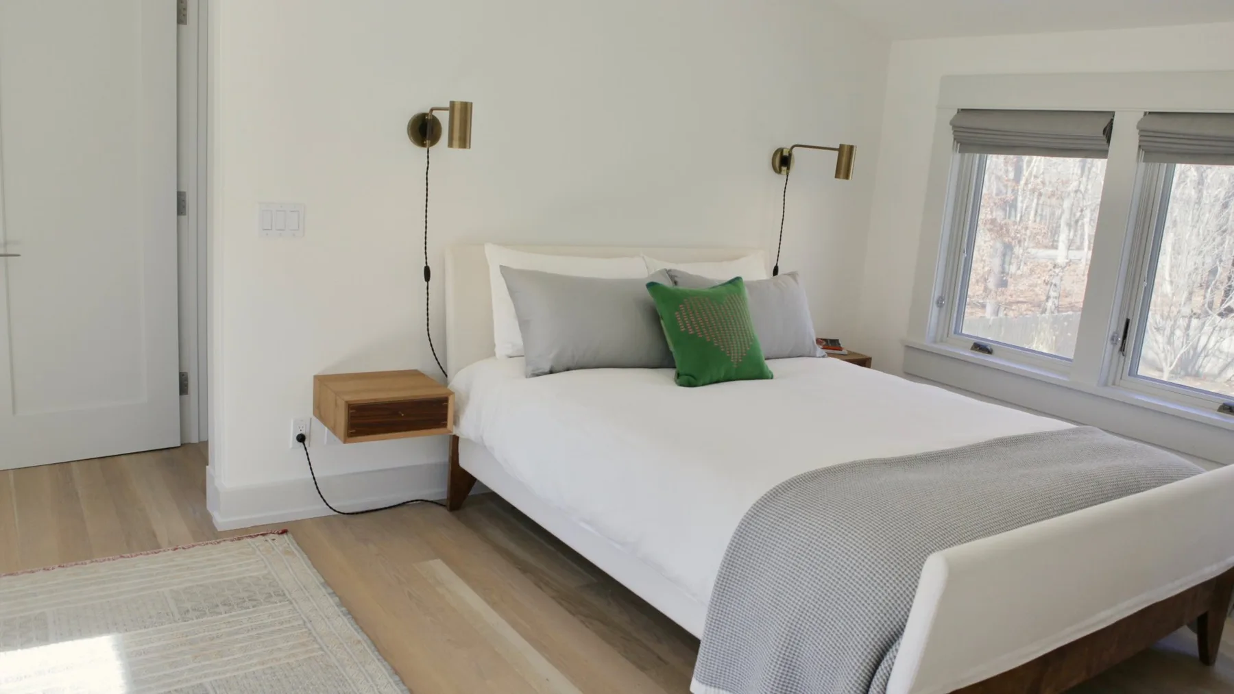 A bright bedroom with a neatly made bed and windows beside it.