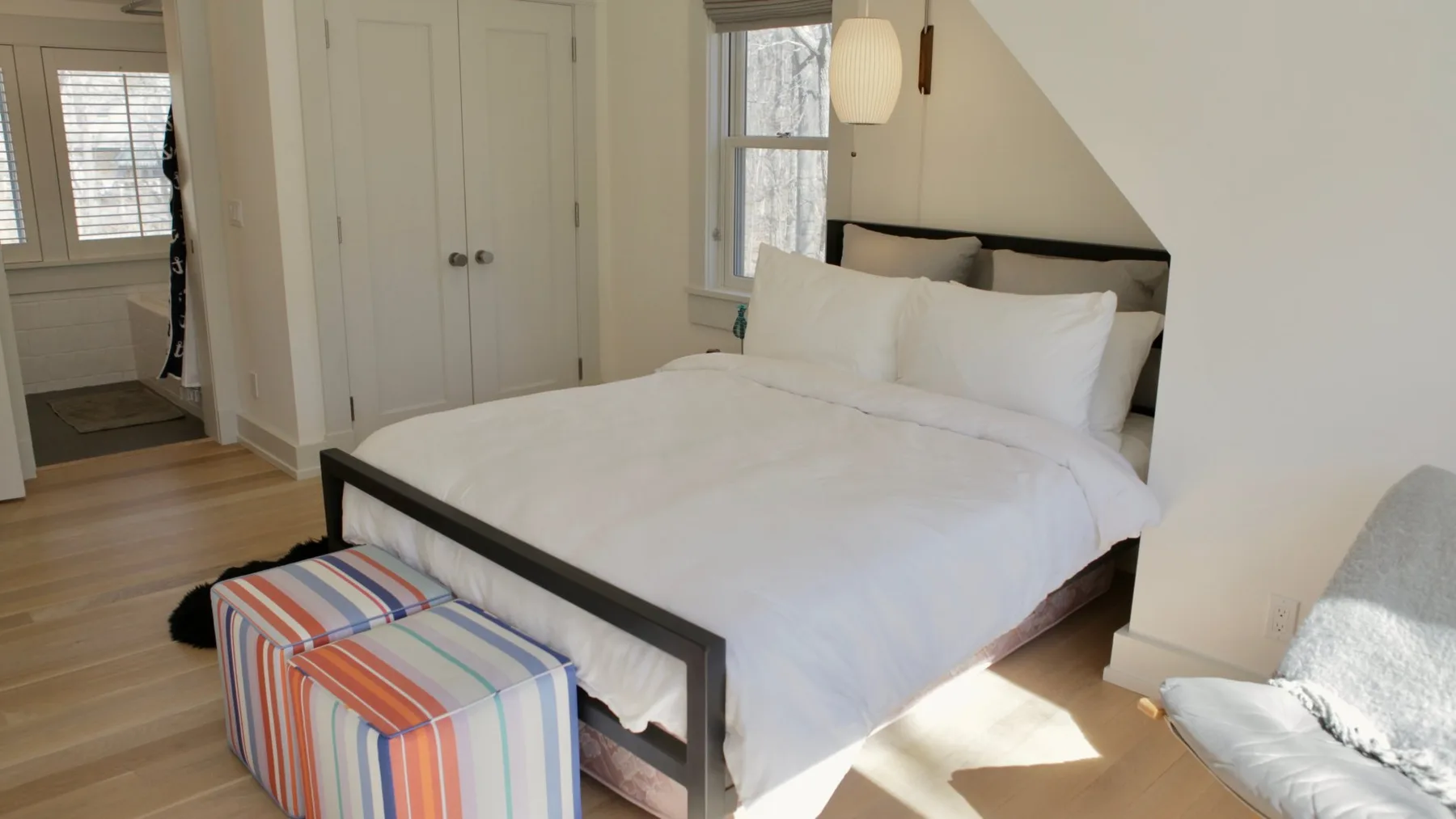 A bedroom with a neatly made bed and a striped ottoman at its foot.