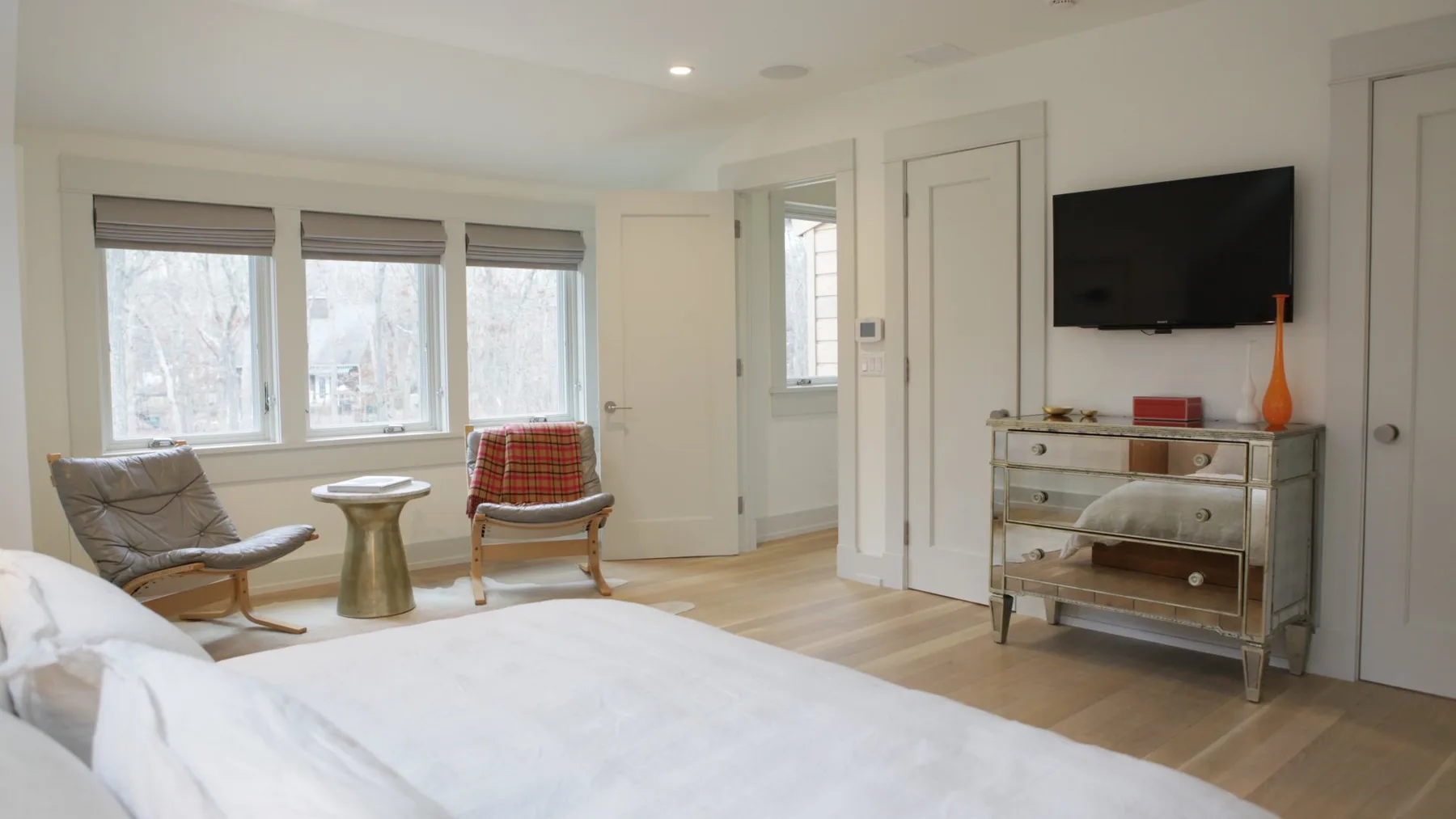A bright bedroom with a bed, seating by the windows, and a wall-mounted television.