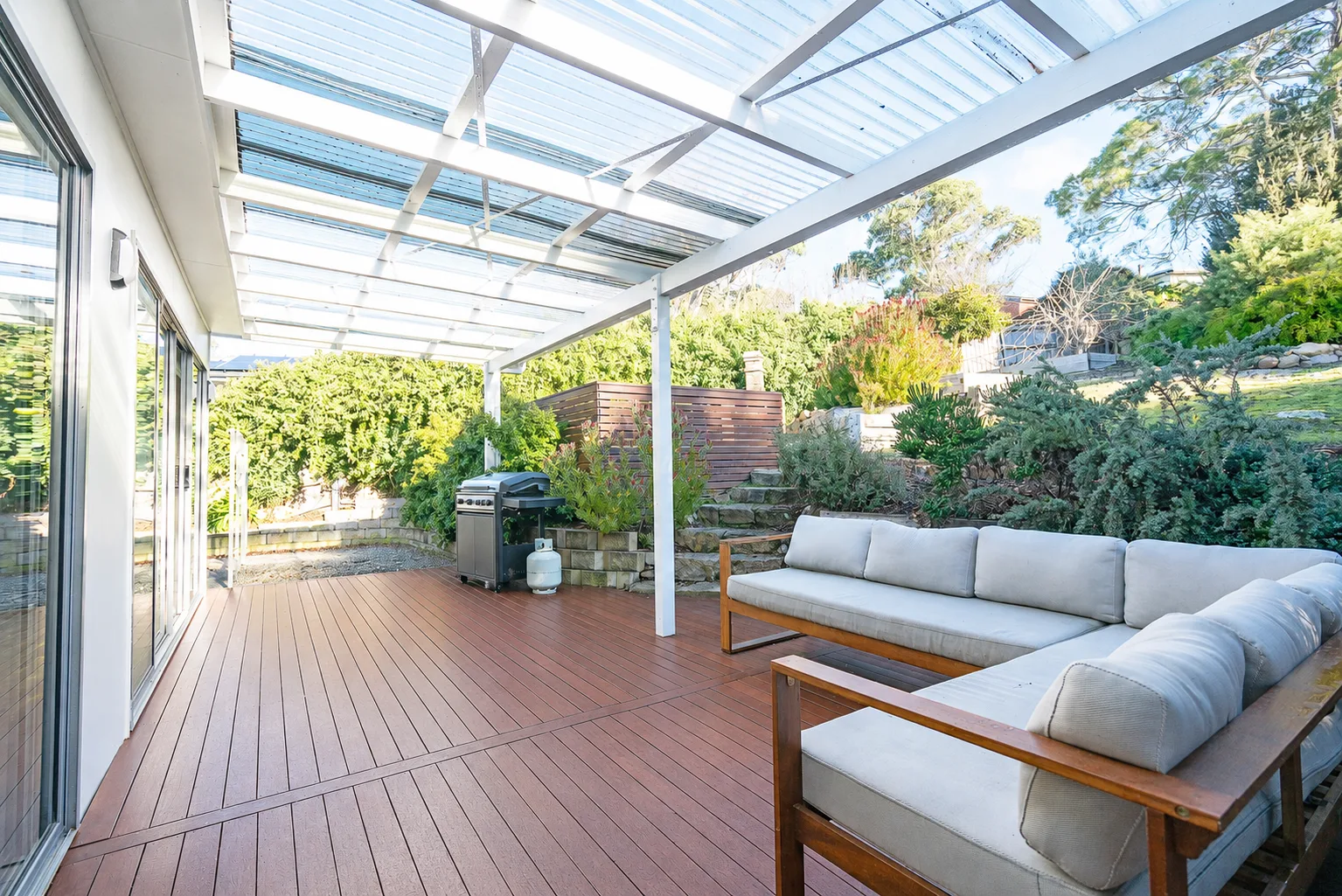A covered outdoor deck has lounge seating and a barbecue beside leafy garden surroundings.