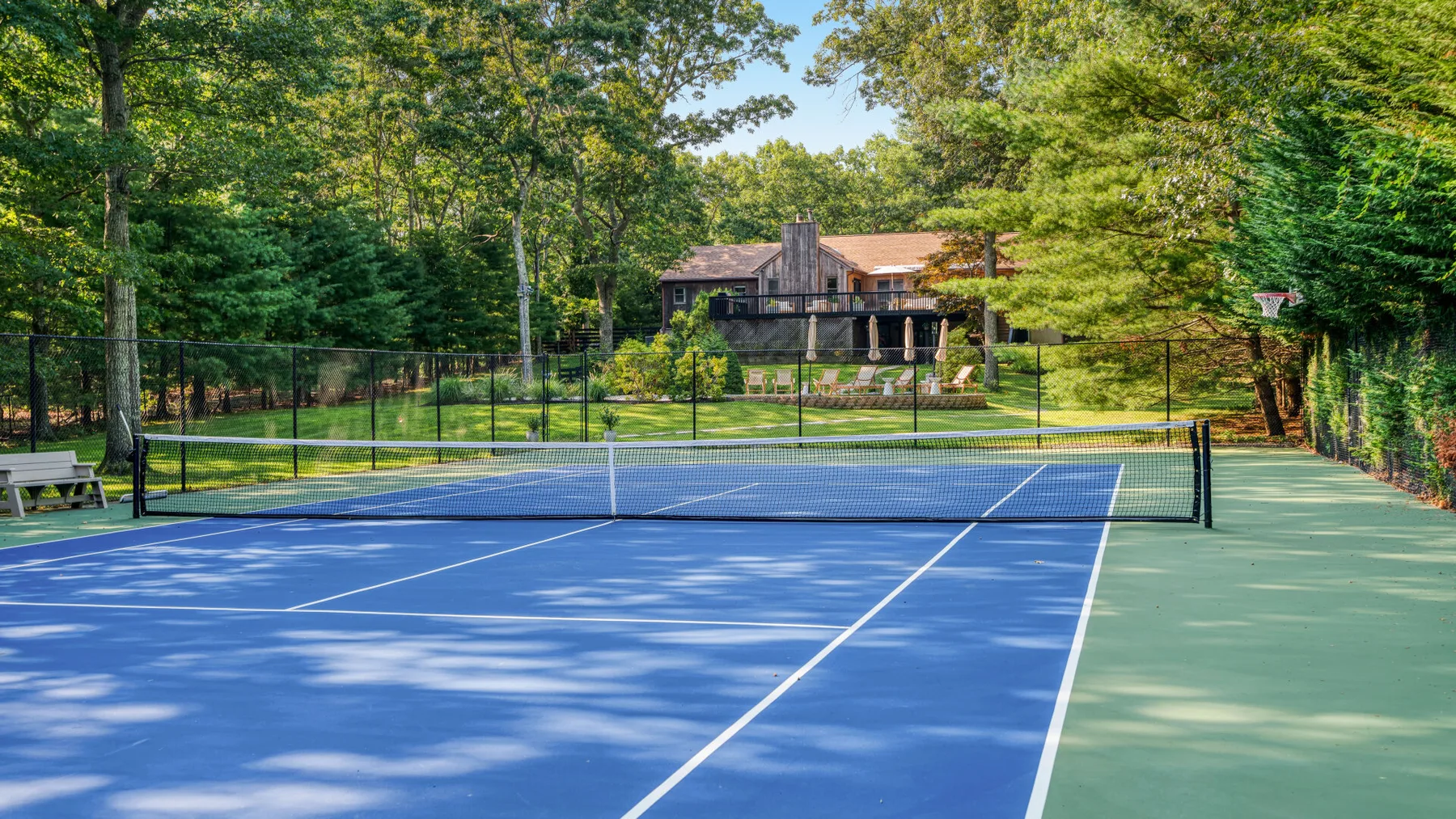 Una cancha de tenis al aire libre está rodeada de árboles, con una casa tras la valla.