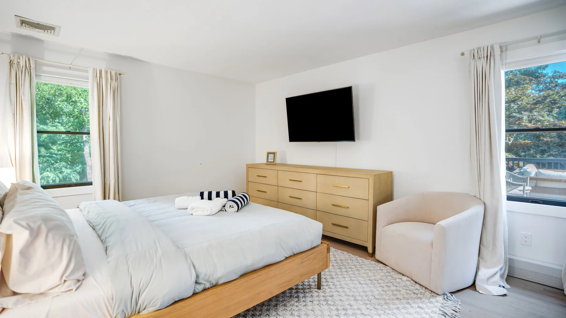 Un dormitorio luminoso con una cama, una cómoda y ventanas a ambos lados.