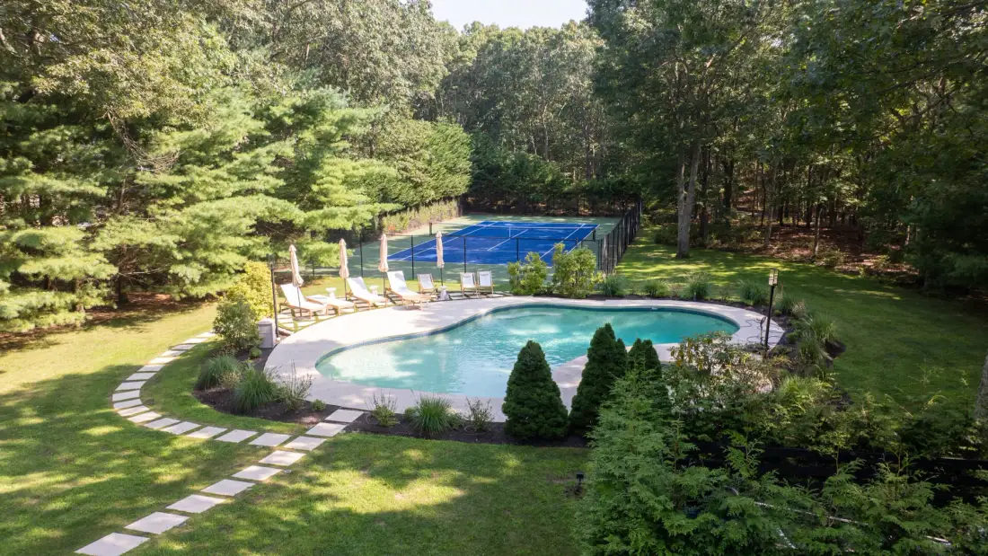 Una piscina al aire libre y una cancha de tenis están rodeadas de jardines y árboles.