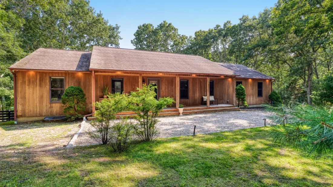 Una casa larga de madera está junto a un camino de grava, rodeada de árboles.