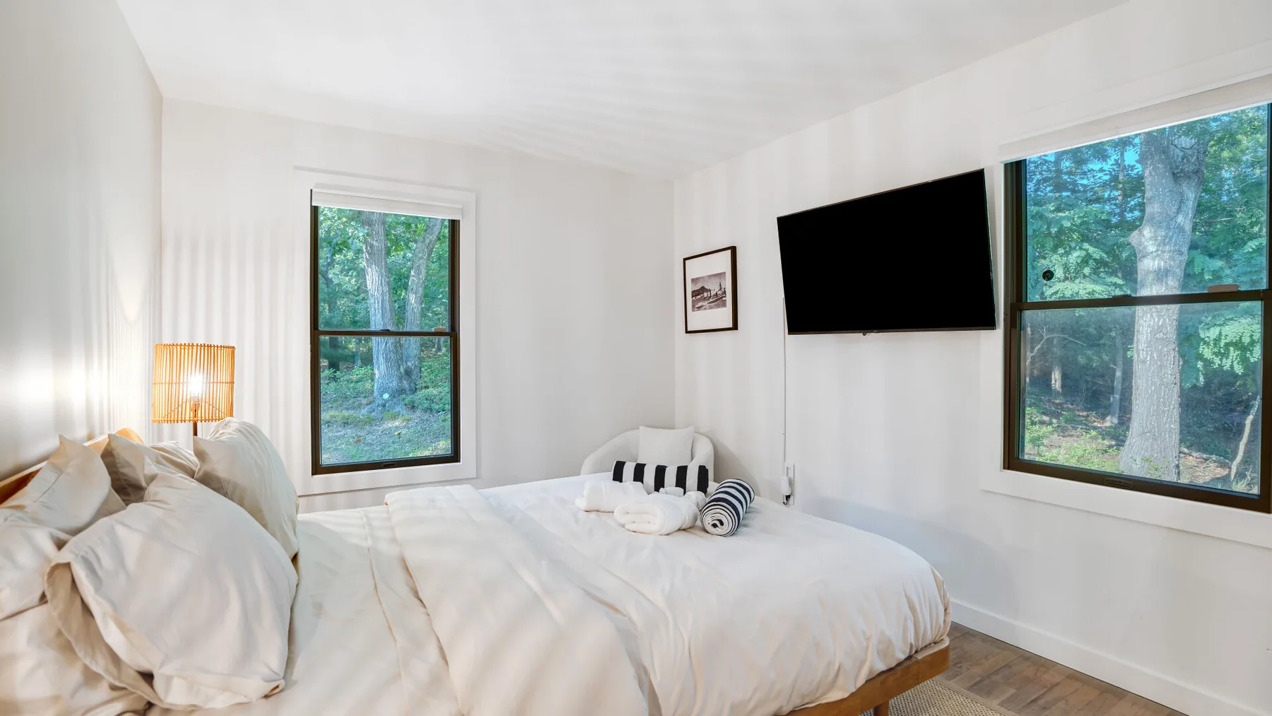 Un dormitorio luminoso con cama, televisor en la pared y ventanas con vista a los árboles.