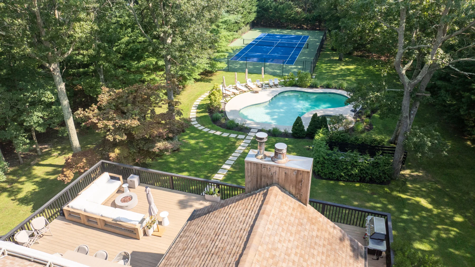 Una vista elevada muestra una terraza de madera, una piscina y una pista de tenis azul rodeadas de árboles.