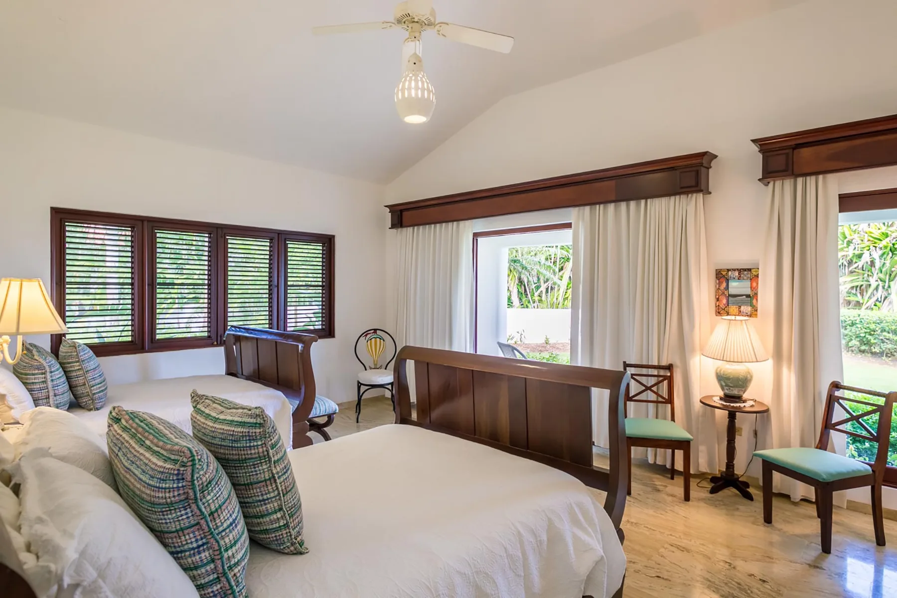 A bright bedroom with a large bed, dark wood furnishings, and windows overlooking greenery.