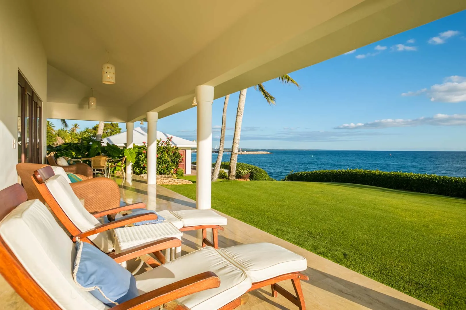 A covered patio with cushioned lounge seating overlooks a green lawn and the ocean.