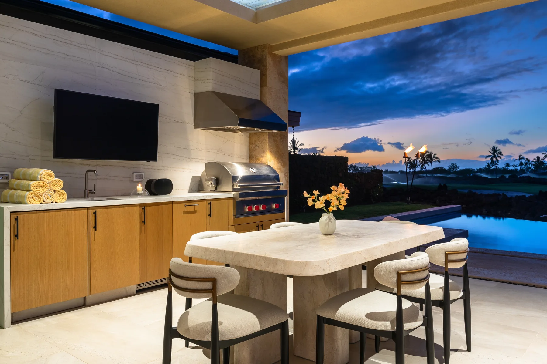 An outdoor kitchen and dining area overlook a pool and sunset sky.