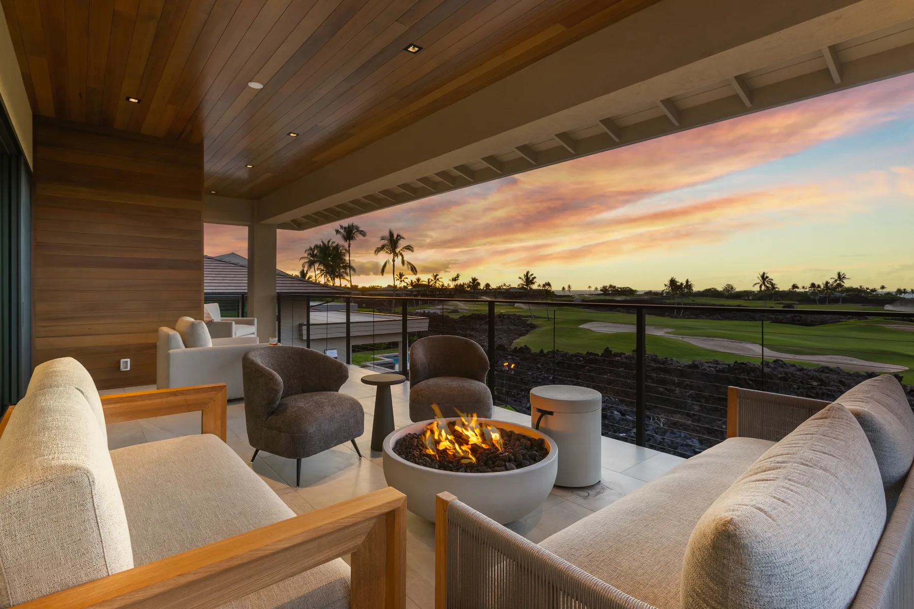 A covered outdoor lounge with seating and a fire pit overlooks a golf course at sunset.