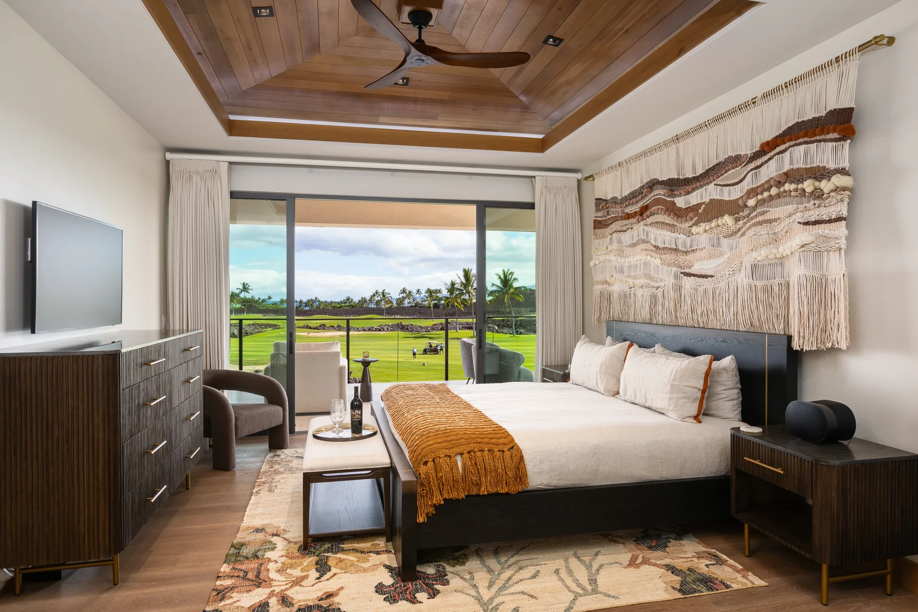A bedroom with a large bed, woven wall hanging, and sliding glass doors overlooking greenery.