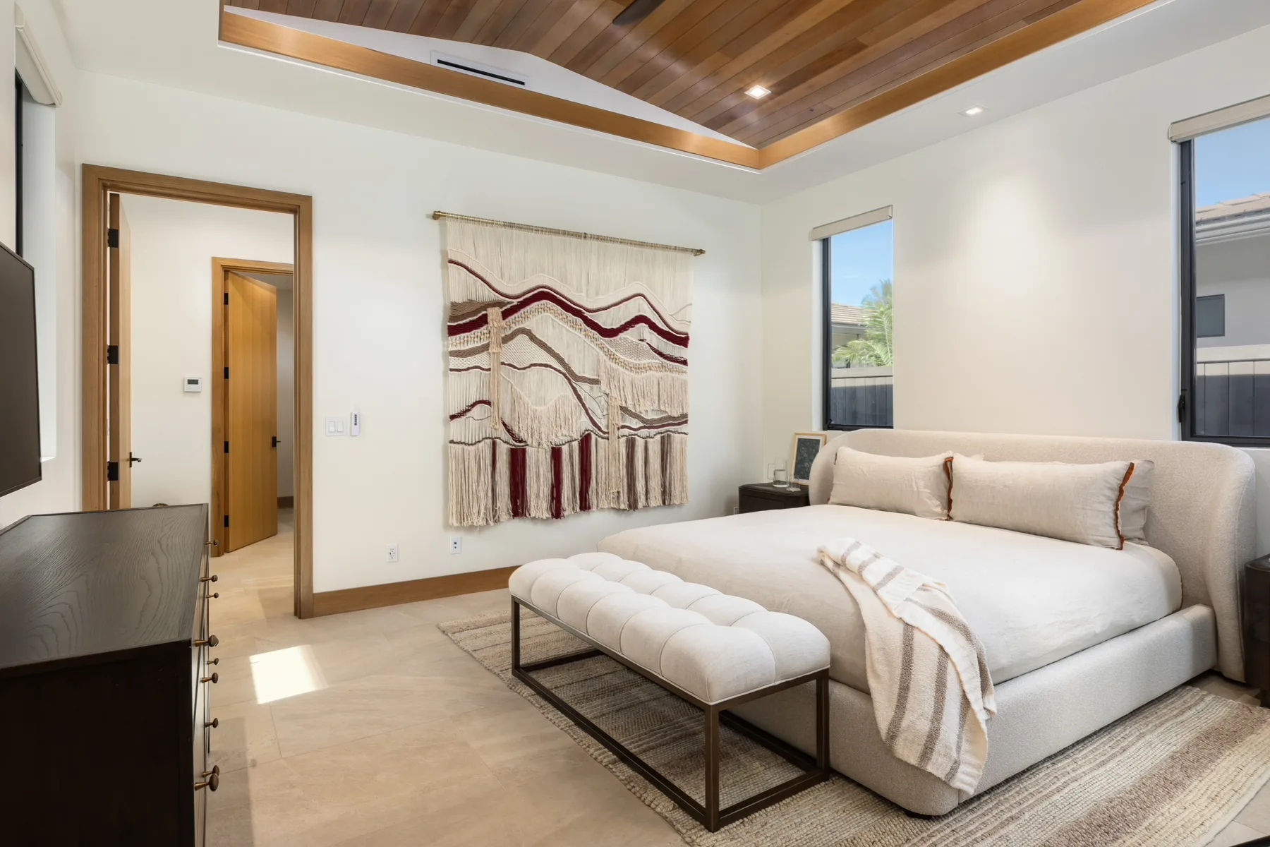 A bright bedroom with a bed, upholstered bench, and patterned wall hanging.