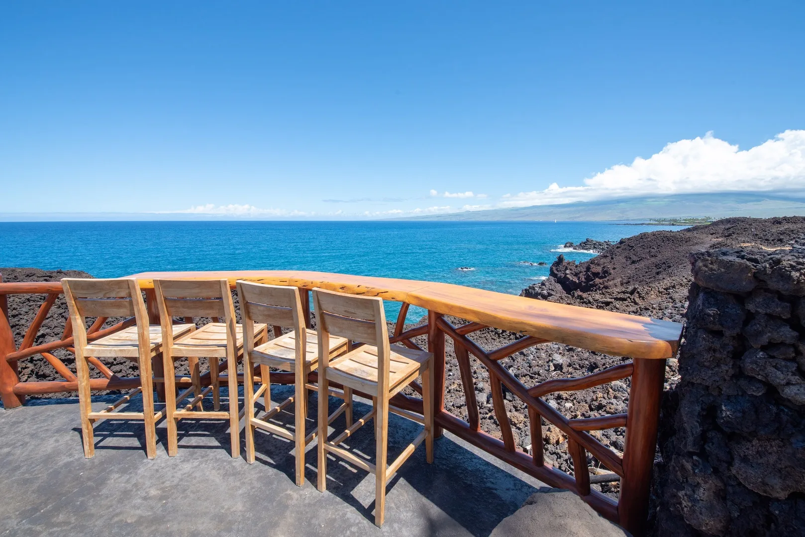 An oceanfront terrace has wooden seating and a railing overlooking the blue water.