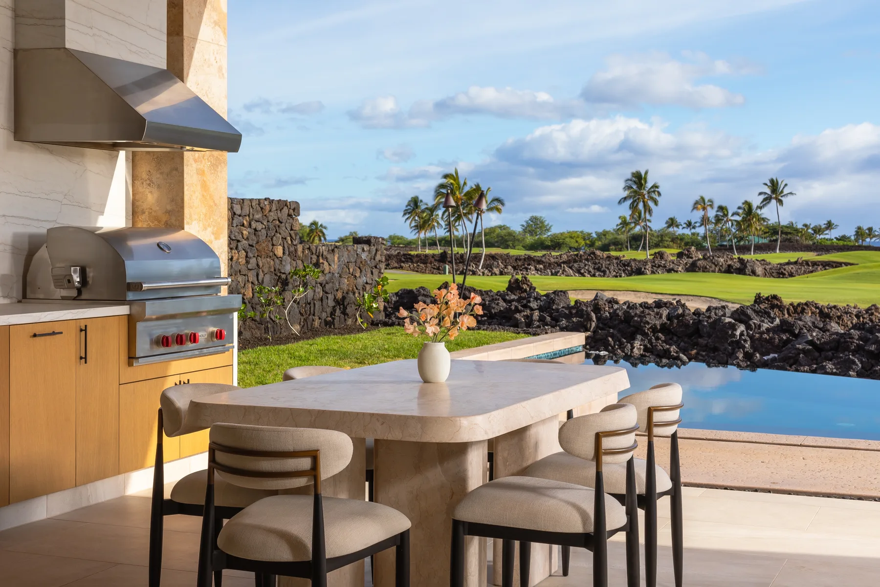 Un comedor al aire libre con parrilla integrada da a una piscina y palmeras.