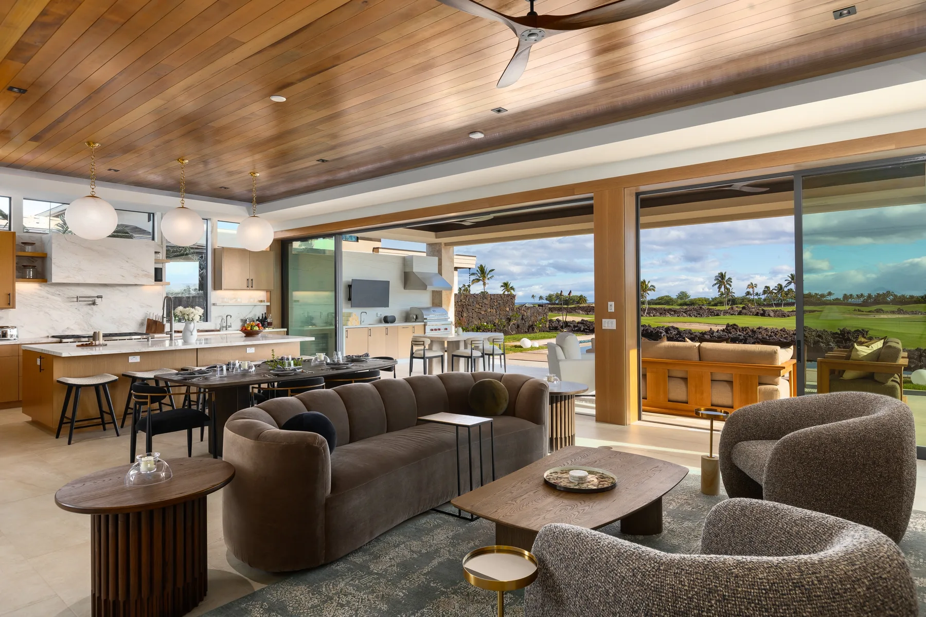 An open-plan living room and kitchen with seating beside wide windows overlooking greenery.