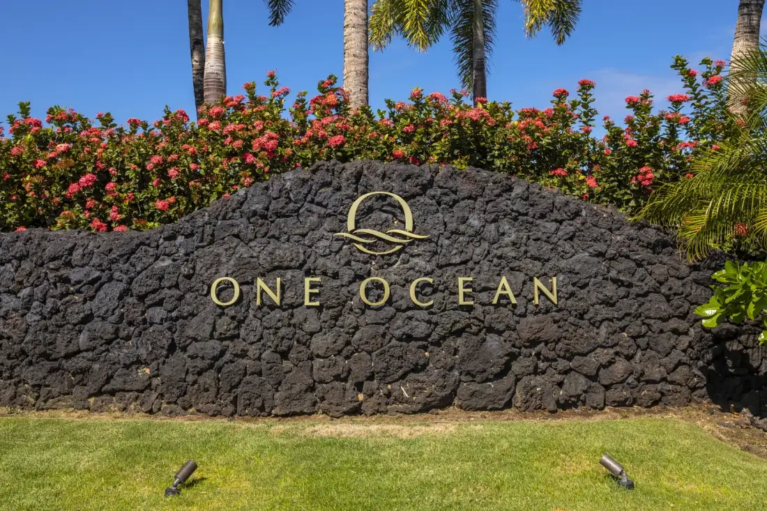 A stone sign reading “One Ocean” stands beneath flowering shrubs and palm trees.