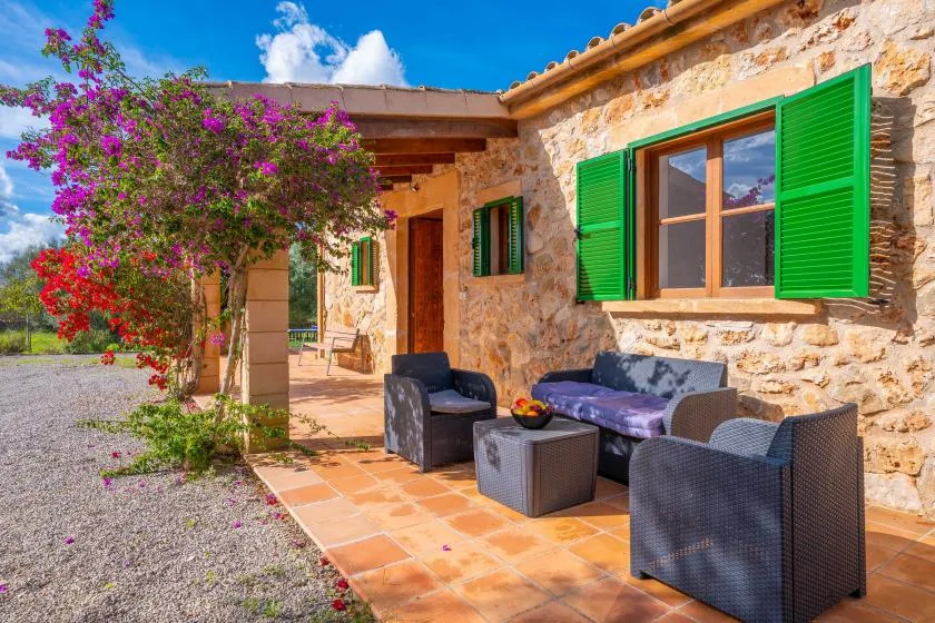 A stone patio with outdoor seating sits beside a house with green shutters.